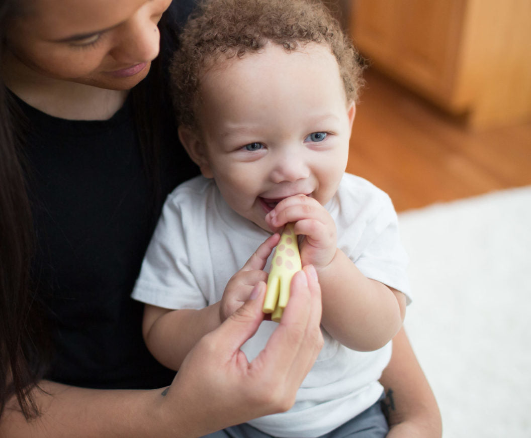 A smiling baby with curly hair, wearing a white shirt, playfully holds a yellow giraffe-shaped toy. The baby sits on a woman's lap, who is gently supporting them. The background shows a cozy home setting with a wooden floor and a white rug.