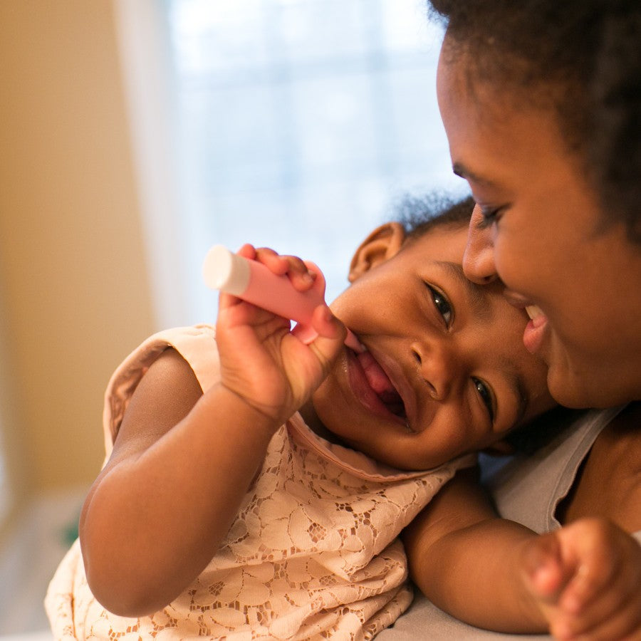 A joyful toddler wearing a lace dress playfully holds a pink tube while being held by an adult. They are smiling and sharing a tender moment, with soft light filtering through a window in the background.