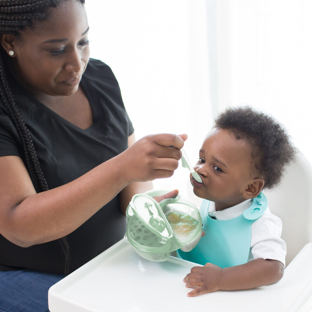 A woman is feeding a baby seated in a high chair. The baby is wearing a teal bib and the woman is holding a container with food and a spoon. The room is bright and airy.
