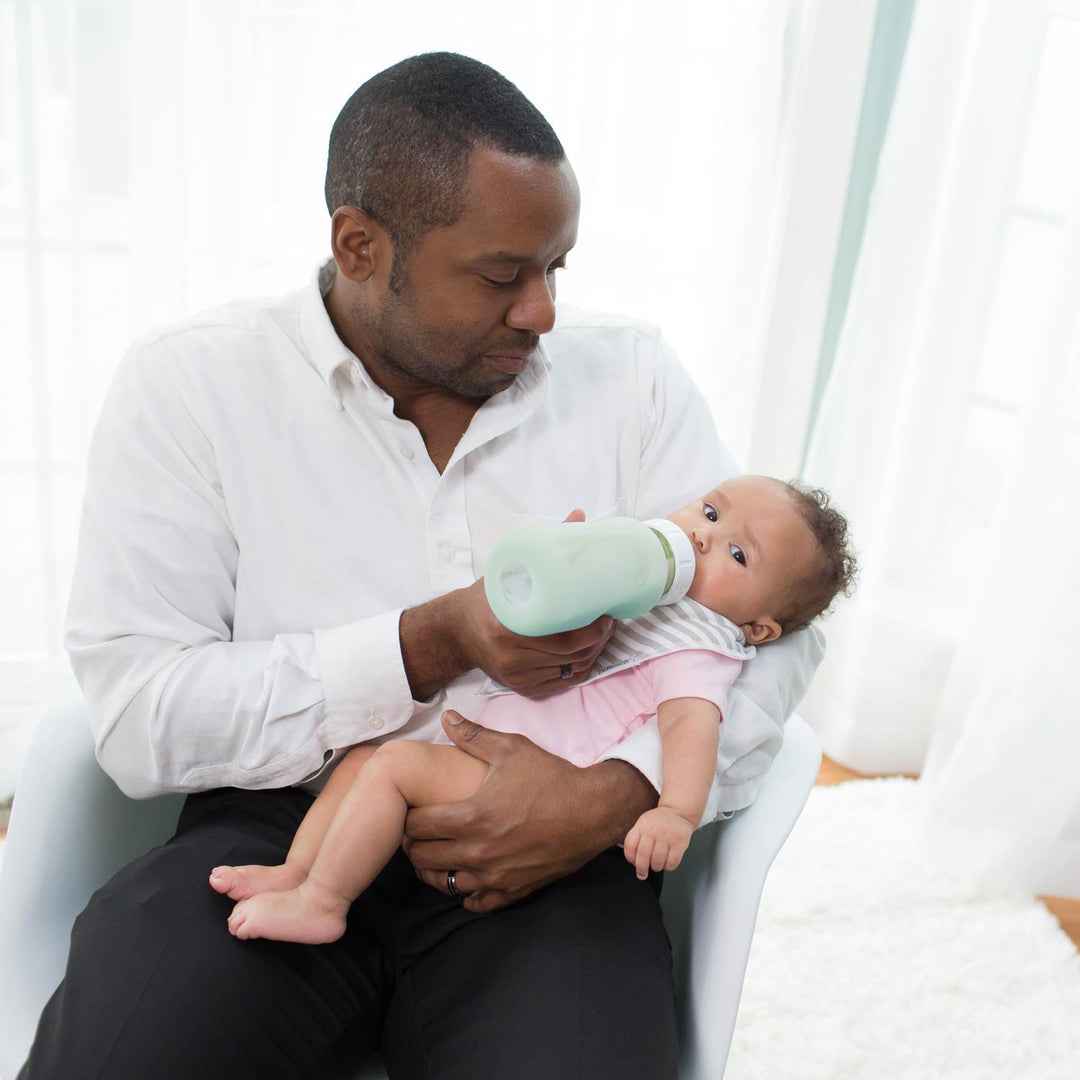 A man in a white button-down shirt and black pants holds a baby wearing a pink outfit while feeding the baby with a Dr. Brown's glass narrow bottle with a mint green sleeve on it in a bright room.
