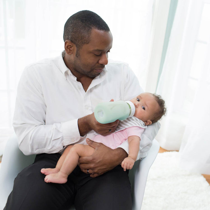 A man in a white button-down shirt and black pants holds a baby wearing a pink outfit while feeding the baby with a Dr. Brown's glass narrow bottle with a mint green sleeve on it in a bright room.