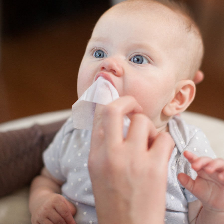 A baby with light hair and blue eyes is sitting up, wearing a gray polka dot onesie. A hand is gently wiping the baby's mouth with a white cloth. The background is softly blurred.