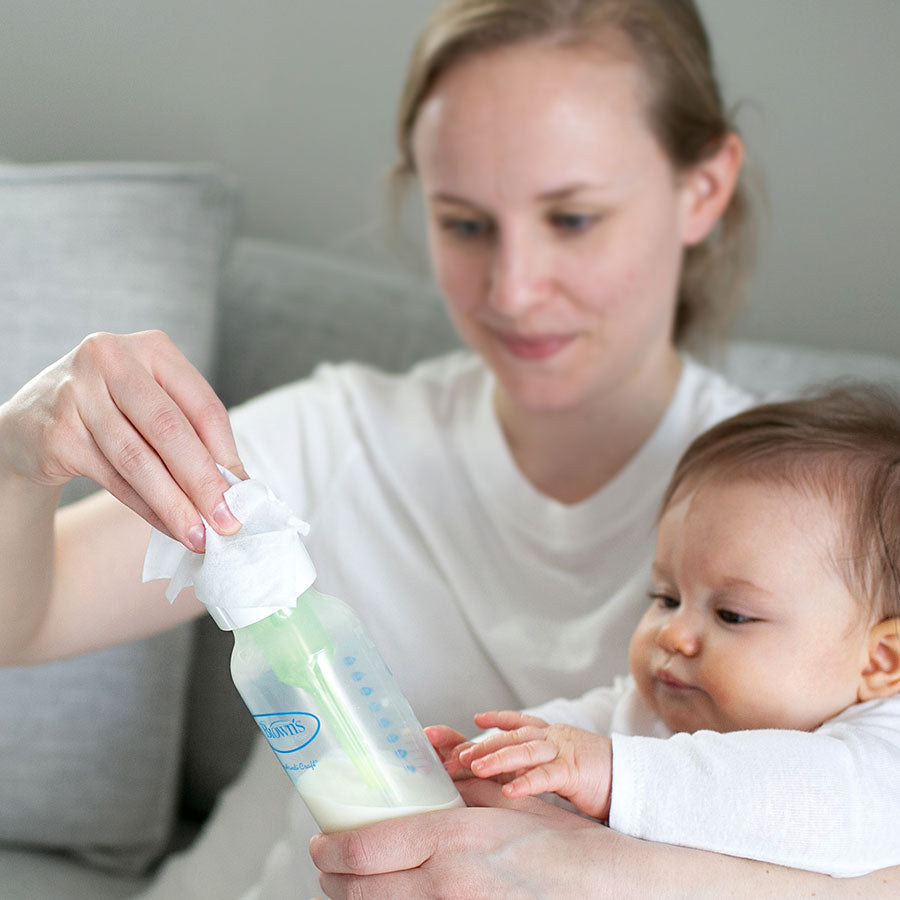 A woman in a white shirt, holding a baby, is cleaning a baby bottle with a tissue. The baby, also dressed in white, reaches out towards the bottle. They're sitting on a couch with a light gray cushion in the background.