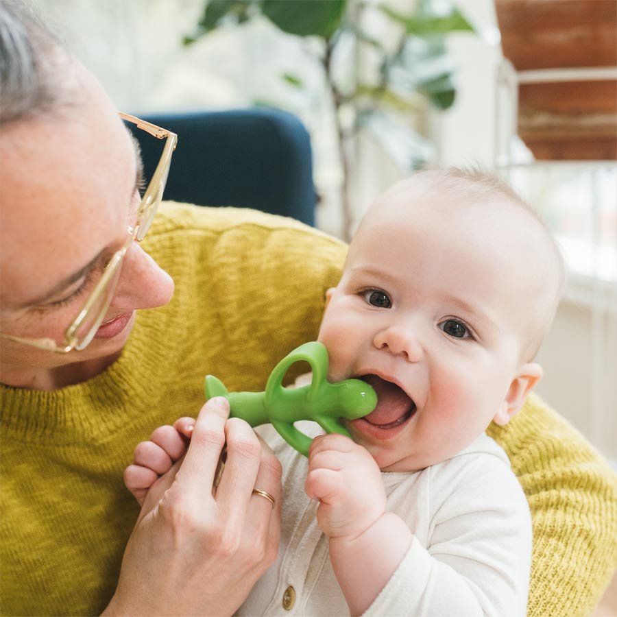 Adult holding a baby wearing a white onesie. The baby is chewing on a green toy with a smile. The adult, wearing glasses and a yellow sweater, is smiling while looking at the baby. Bright, cozy setting with plants in the background.