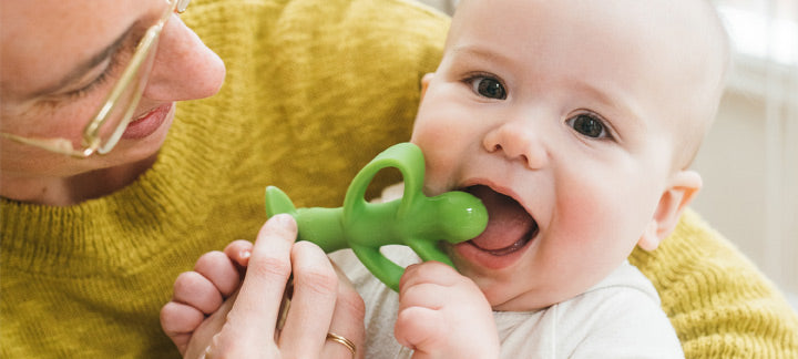 Baby holding a green teething toy with a person in a yellow sweater