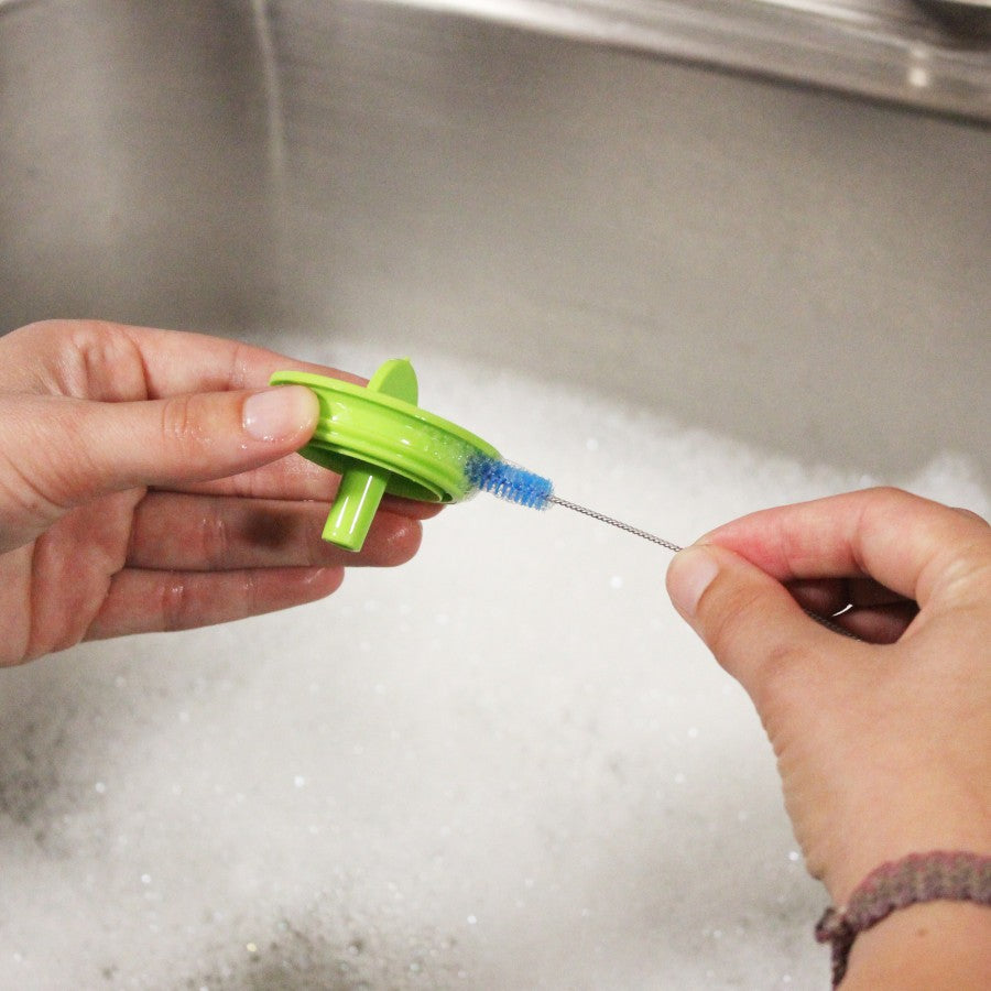 Two hands are cleaning a green plastic bottle part with a small blue brush over a sink filled with soapy water.