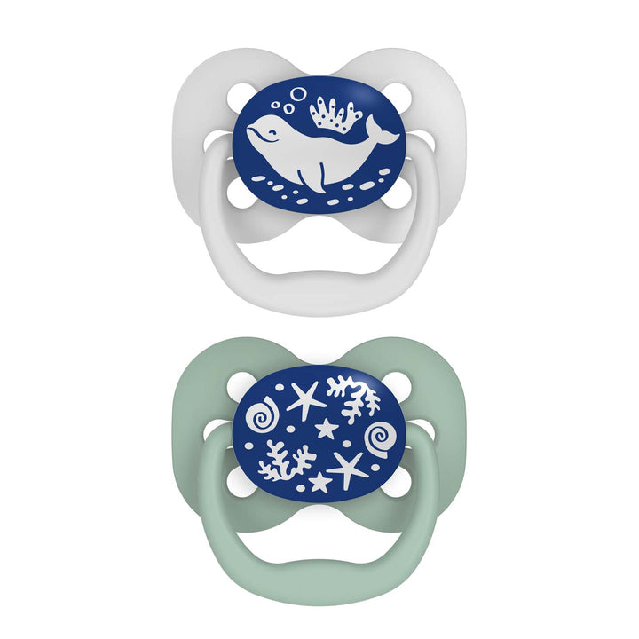 Dr. Brown's™ Advantage™ Glow-in-the-Dark Pacifiers, 2 Count, feature nautical designs: a white with blue shield and whale pacifier, and a light green with a blue shield displaying seashells and starfish pacifier. These symmetrical soothers ensure your baby's comfort.