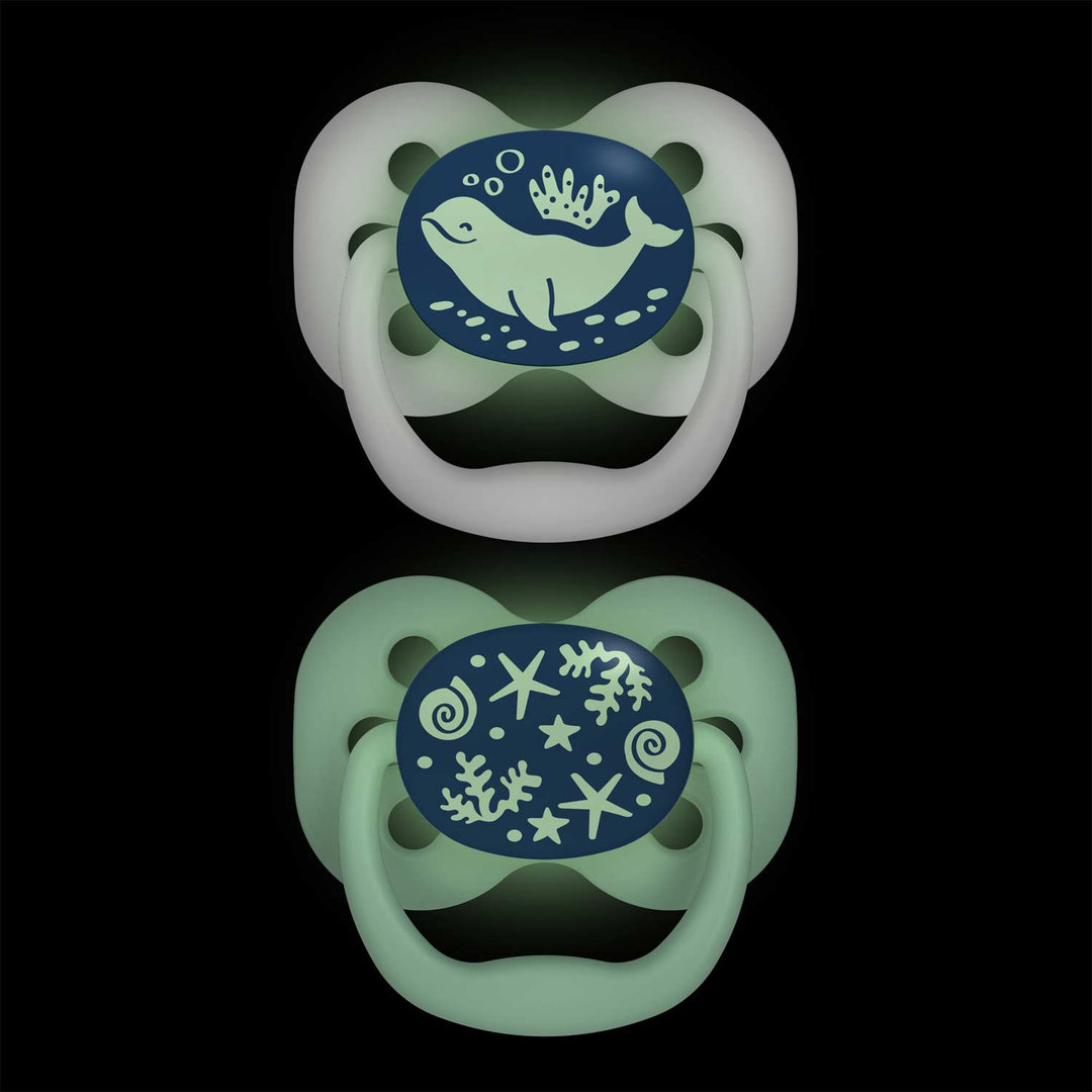 Dr. Brown's™ Advantage™ Glow-in-the-Dark Pacifiers, 2 Count, feature two reversible pacifiers with glowing designs on black. The top soother showcases a whale with bubbles and seaweed, while the bottom glows in the dark with ocean motifs like shells and coral, both having a greenish hue.