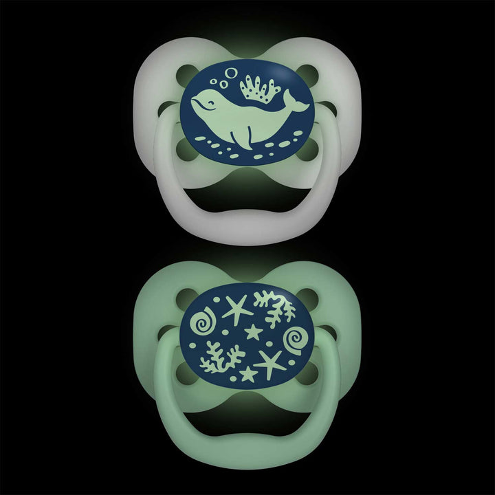 Dr. Brown's™ Advantage™ Glow-in-the-Dark Pacifiers, 2 Count, feature two reversible pacifiers with glowing designs on black. The top soother showcases a whale with bubbles and seaweed, while the bottom glows in the dark with ocean motifs like shells and coral, both having a greenish hue.