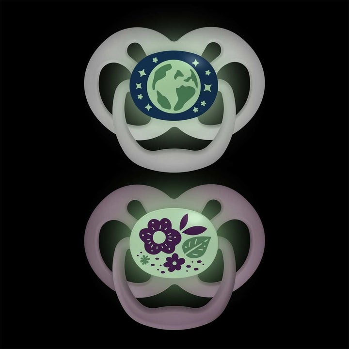 On a black background, Dr. Brown's™ Advantage™ Glow-in-the-Dark Pacifiers feature one with an Earth and stars design on blue-green, and another with purple flowers and green leaves. Both include convenient reversible bulbs.