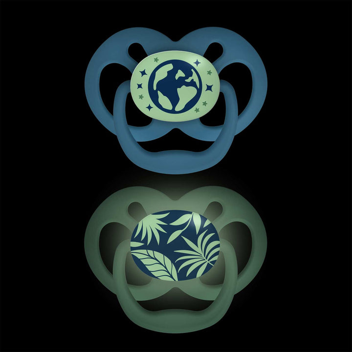 Dr. Brown's™ Advantage™ Glow-in-the-Dark Pacifiers, 2 Count, features two attention-grabbing designs on a black background: a glowing globe motif with stars and a symmetrical dark green soother adorned with green leaves.