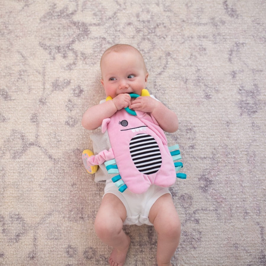 A baby lying on a soft, light-colored carpet, playfully chewing on a pink and blue striped stuffed toy. The baby is wearing a white diaper and looking upward with a curious expression.