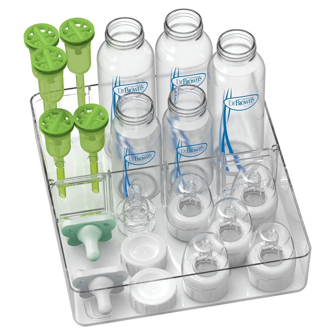 A clear plastic organizer tray holding multiple white and clear baby bottles, pacifiers, and bright green bottle-top accessories, photographed on a white background. Several bottles show the Dr. Brown's brand name.