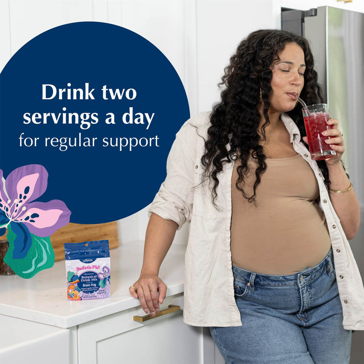 A pregnant woman with curly dark hair stands in a bright kitchen, drinking from a glass of red liquid. On the counter beside her is a small package of dietary supplement powder, and a large blue graphic overlay reads, 'Drink two servings a day for regular support.