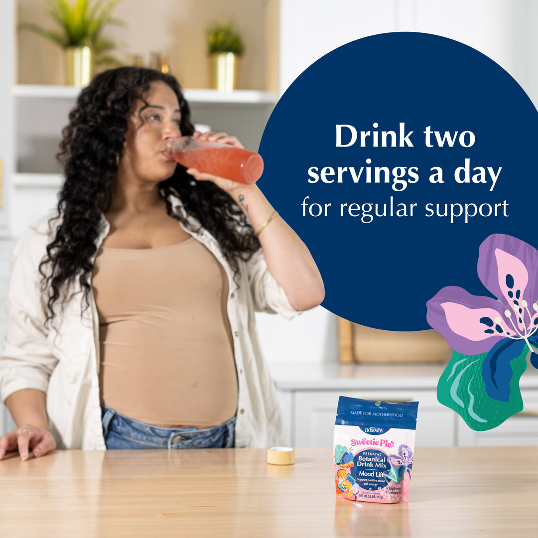 A pregnant woman in a kitchen setting drinks a pink beverage from a glass, with a package of Prenatal Probiotics on the table in front of her. A large dark blue text bubble reads 'Drink two servings a day for regular support' with a purple and green flower graphic on the right.
