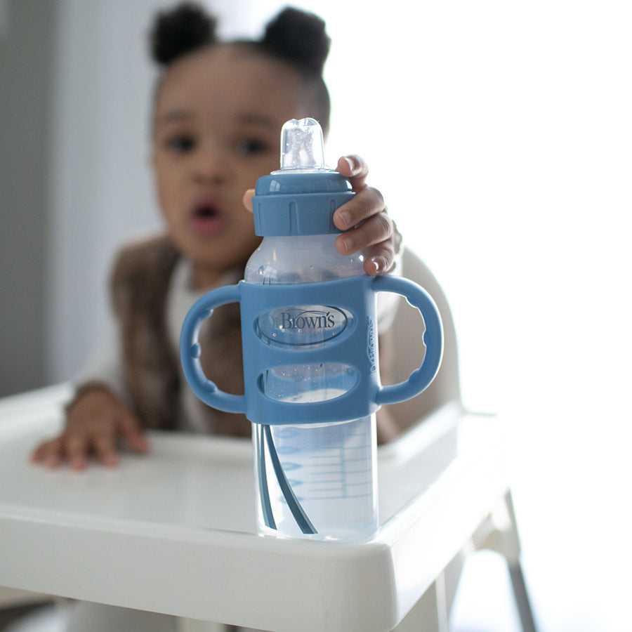 A toddler sitting in a high chair reaches out for a blue baby bottle with handles. The bottle is placed on the tray of the chair. The focus is on the bottle, which is closer to the camera.