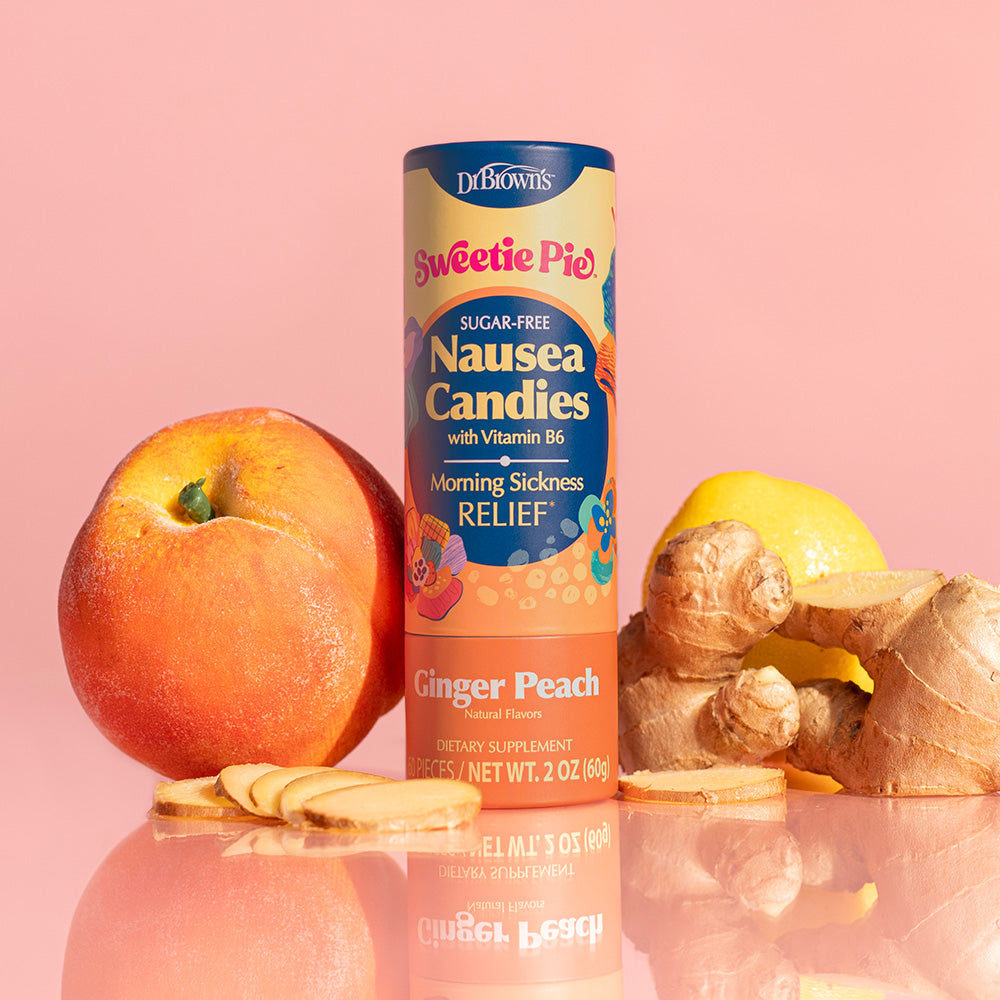 A bottle of Dr. Brown's Sweetie Pie Ginger Peach Nausea Candies stands upright on a reflective pink surface, surrounded by a peach, ginger root slices, and a lemon against a solid pink background.