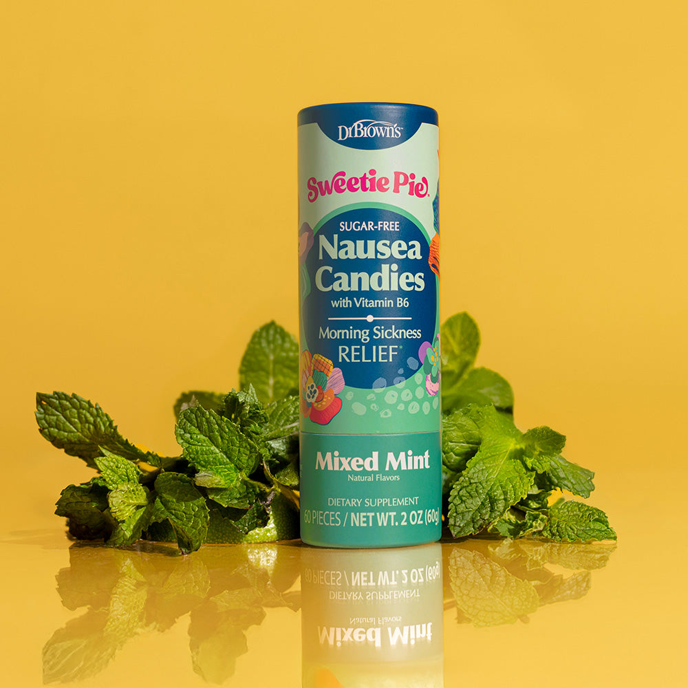 A bottle of Dr. Brandes Sweetie Pie Nausea Candies in Mixed Mint flavor, styled with fresh mint leaves on a reflective yellow surface against a solid yellow background.