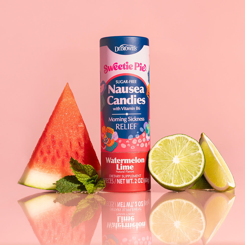 A Dr. Brown's Sweetie Pie Nausea Candies tube packaging stands upright on a reflective pink surface, flanked by a watermelon slice, mint leaves, and lime wedges against a solid pink background.