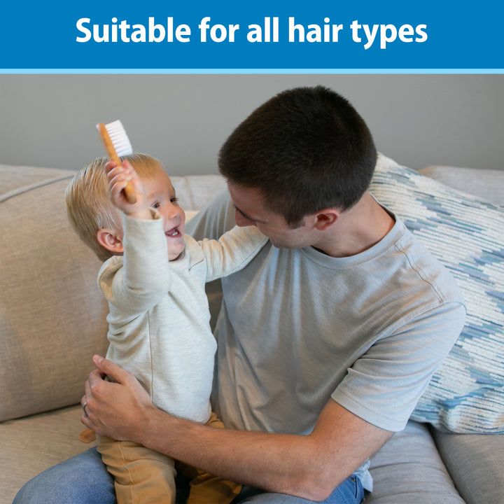 A smiling young child holds Dr. Brown's Soft and Safe Baby Brush while sitting on an adult's lap on a couch, with the adult looking fondly at the child. Text at the top says, "Suitable for all hair types."