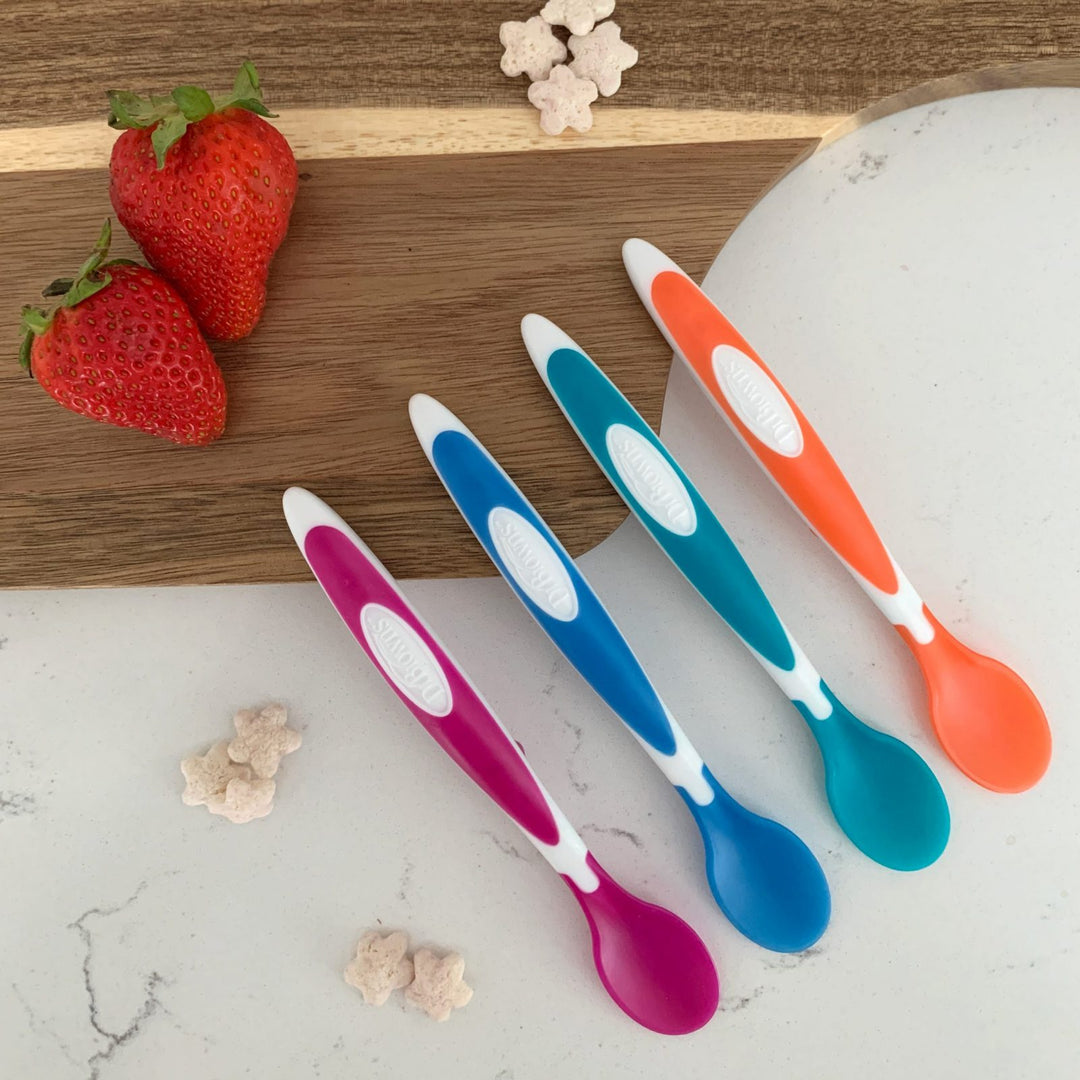 Four colorful baby spoons—pink, blue, teal, and orange—are placed on a wooden and white surface. Three strawberries and small, star-shaped cereal pieces are nearby.