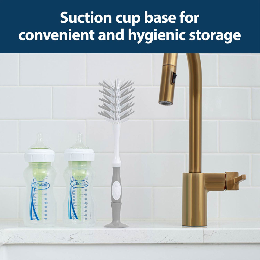 The Dr. Brown’s™ Deluxe Bottle Brush with a suction cup base stands upright for easy cleaning beside two baby bottles and a brass kitchen faucet on a white countertop.