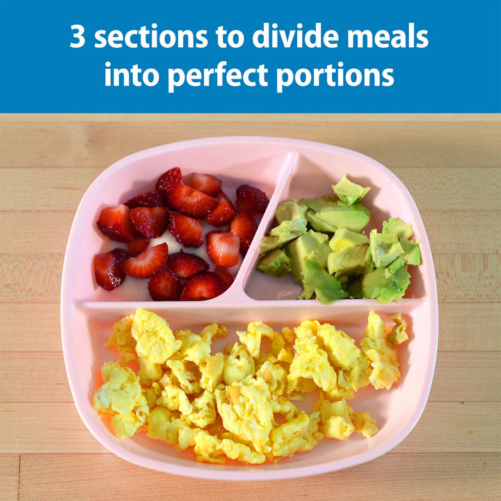 A pink No-Slip Divided Suction Plate on a light wood surface, filled with sliced strawberries, avocado pieces, and scrambled eggs. A blue banner at the top reads, '3 sections to divide meals into perfect portions.