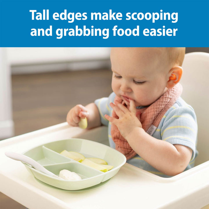 A baby seated in a high chair using a mint No-Slip Divided Suction Plate with a spoon on the tray. A blue banner at the top reads, 'Tall edges make scooping and grabbing food easier.
