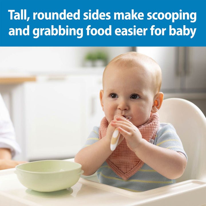 A baby sitting in a high chair wearing a pink bib and holding a white spoon, with a mint colored suction bowl on the tray. A blue text banner at the top reads, 'Tall, rounded sides make scooping and grabbing food easier for baby.