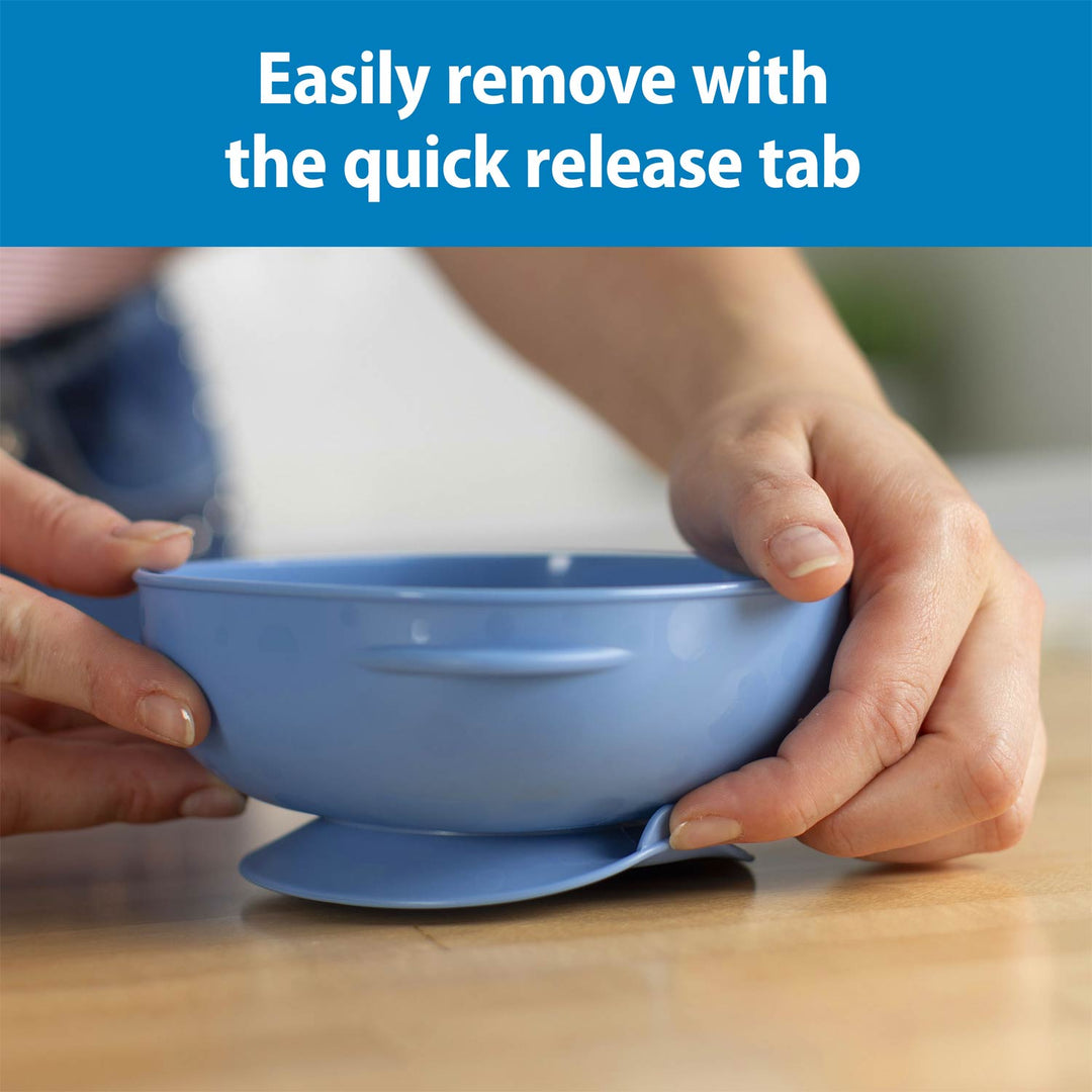 A blue silicone bowl with a suction base is being held by two hands over a wooden table, with a blue text banner at the top reading 'Easily remove with the quick release tab.'.