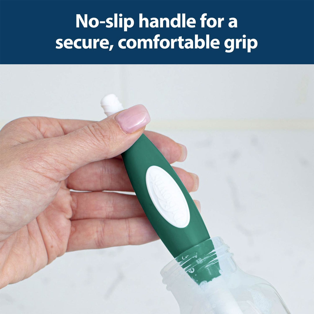 A hand holds a green and white brush with a no-slip handle, cleaning the inside of a clear bottle against a light background.
