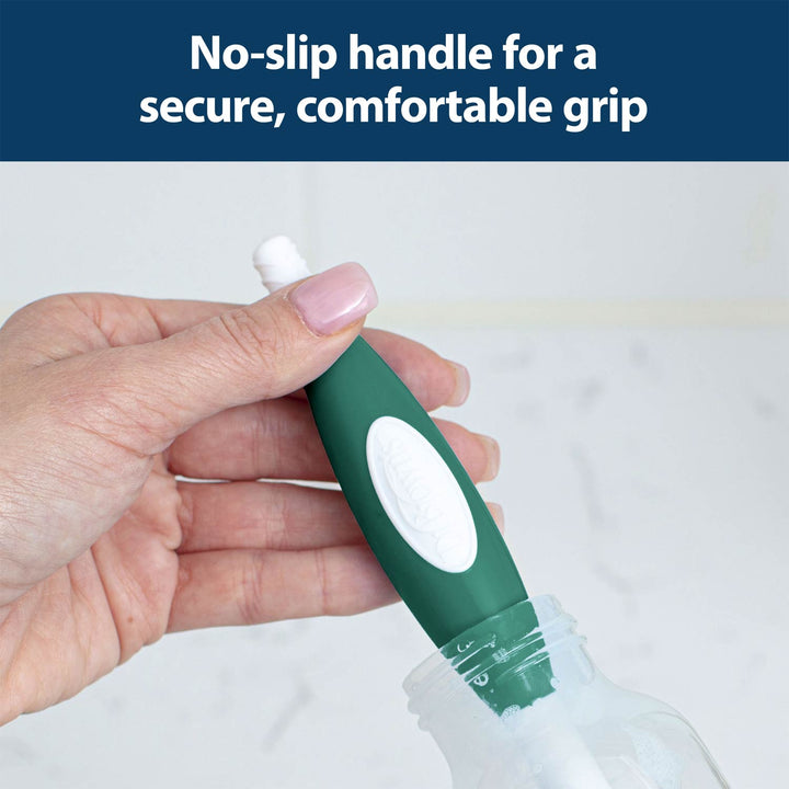 A hand holds a green and white brush with a no-slip handle, cleaning the inside of a clear bottle against a light background.