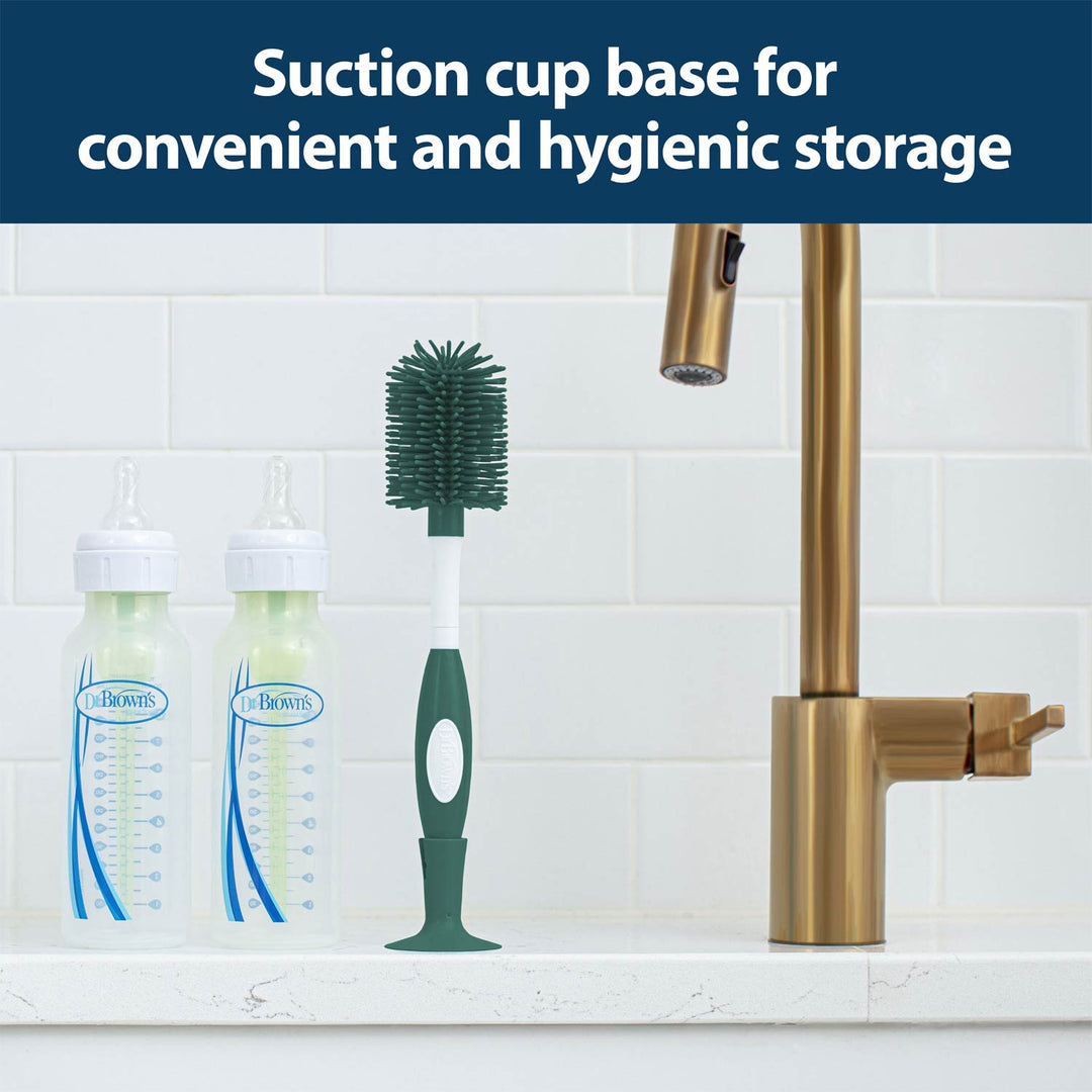 A bottle brush with a suction cup base stands upright on a kitchen counter next to two baby bottles and a brass faucet.