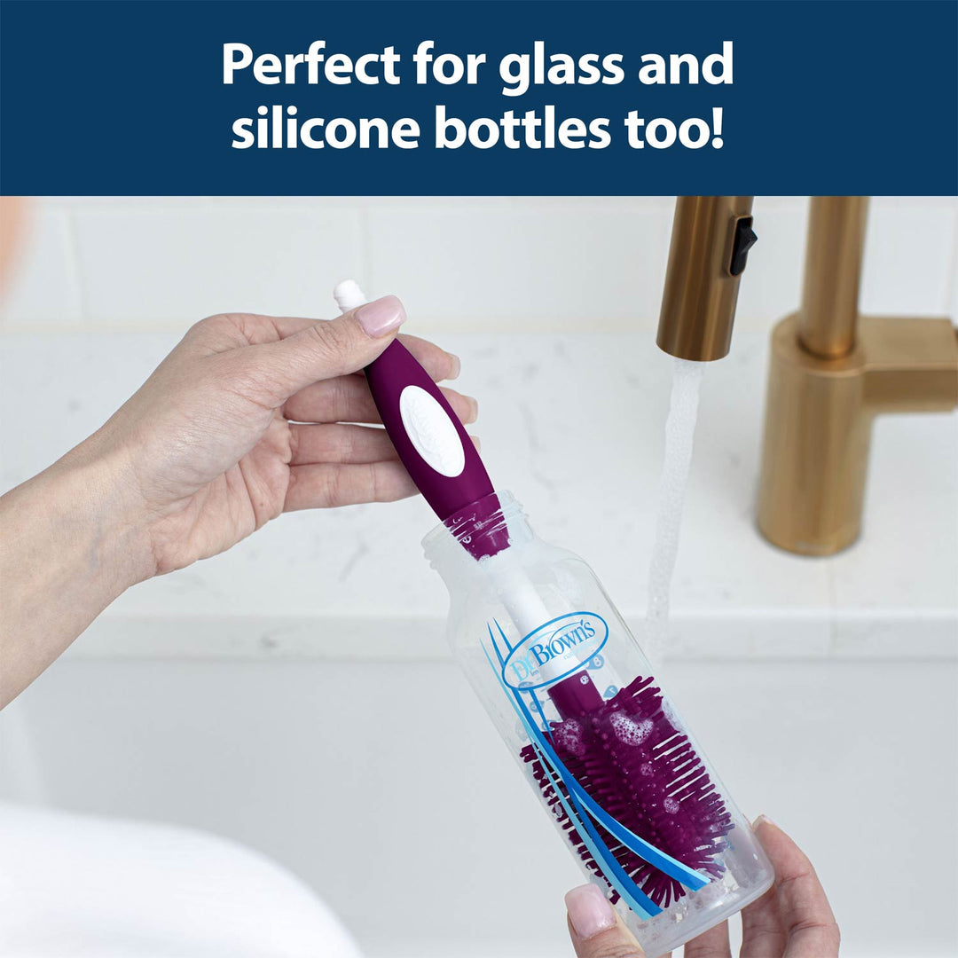 A person holds a dark pink and white cleaning brush inside a clear bottle under running water at a sink, with a dark blue banner at the top reading 'Perfect for glass and silicone bottles too!'.