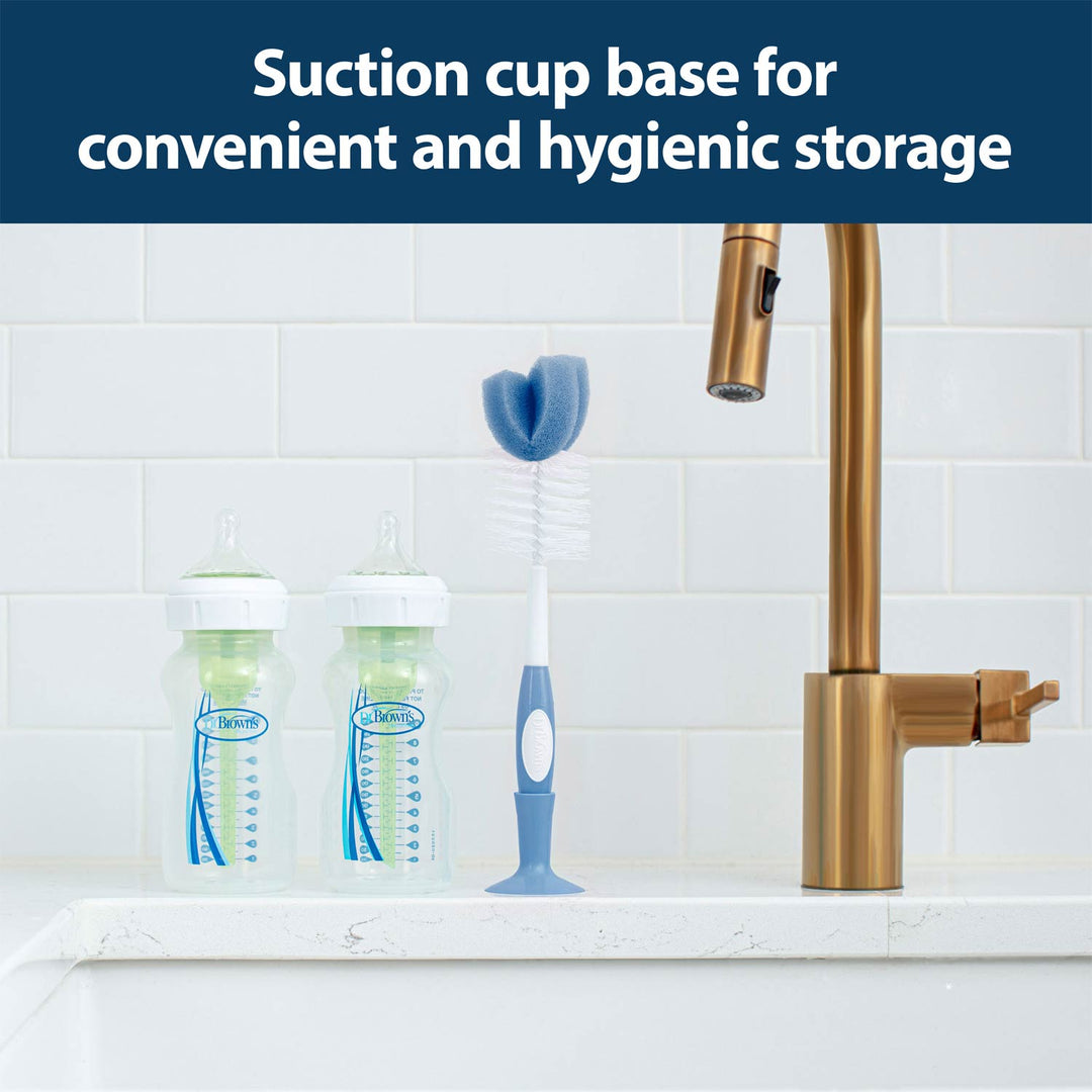 Dr. Brown's™ Baby Bottle Brush with nipple cleaner stands upright on a kitchen counter by two baby bottles and a sink; text reads: Suction cup base for convenient, hygienic storage.