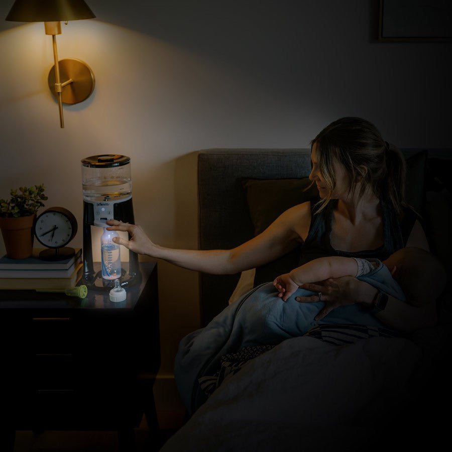 In a dimly lit room, a woman is seated on a bed, feeding her baby. Next to her is the Dr. Brown’s™ Insta-Prep Warm Water Dispenser, providing warm milk for her little one. يوجد مصباح على الحائط وساعة بجانبه.