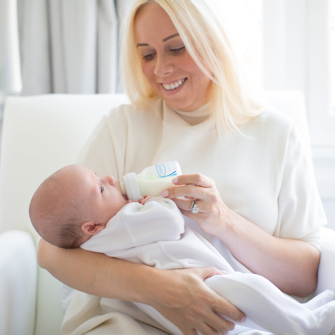 A smiling woman with blonde hair is seated on a white chair, bottle-feeding an infant dressed in a white outfit using Dr. Brown's Natural Flow® Options+™ Wide-Neck Baby Bottle Replacement Vent Kit, 9oz, 2-Pack. The room is softly lit, creating a warm and nurturing atmosphere.