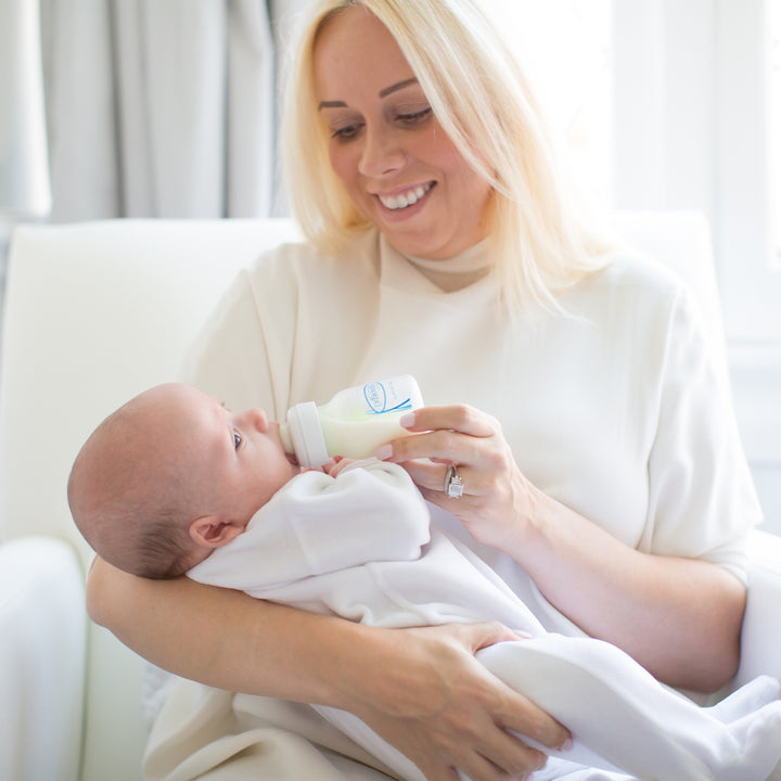 A smiling woman with blonde hair is seated on a white chair, bottle-feeding an infant dressed in a white outfit using Dr. Brown's Natural Flow® Options+™ Wide-Neck Baby Bottle Replacement Vent Kit, 9oz, 2-Pack. The room is softly lit, creating a warm and nurturing atmosphere.