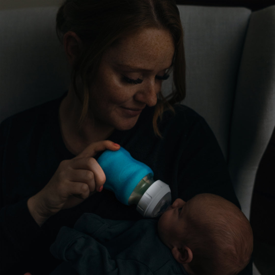 A woman with red hair gently feeds a baby using a Dr. Brown's Natural Flow® Options+™ Wide-Neck Glass Bottle Silicone Sleeves while seated on a chair. The scene is intimate, enhanced by soft lighting and a serene atmosphere.