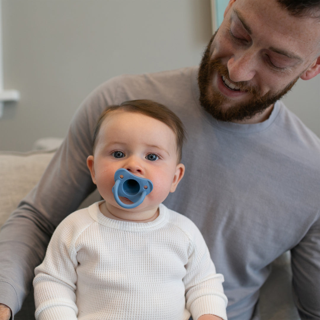 A baby with a blue Dr. Brown’s® HappyPaci™ 100% Silicone One-Piece Pacifier sits on an adult's lap. The adult, dressed in a gray shirt, smiles as they gaze down at the baby, who is wearing a white shirt. They are seated on a couch in a gently illuminated room.