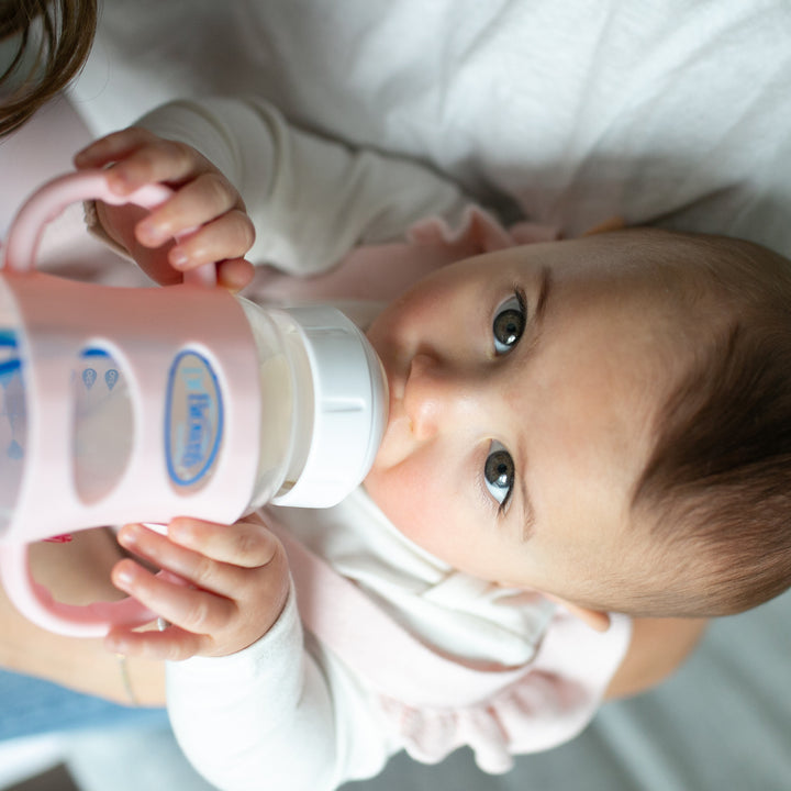 A baby with dark hair, wearing a white shirt, is lying down while independently drinking from a pink and white Dr. Brown’s® Milestones™ Wide-Neck Silicone Handled bottle by Dr. Brown's, looking upwards and showing off their independent drinking skills with a soft blanket in the background.