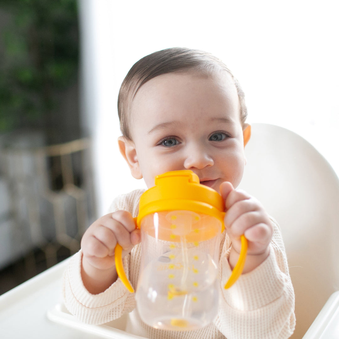 A baby with light skin and dark hair sits in a high chair, happily holding a Dr. Brown's™ Milestones™ Baby’s First Straw Cup, featuring its spill-proof lid, while smiling at the camera. The background is softly blurred, creating a bright and cozy atmosphere.