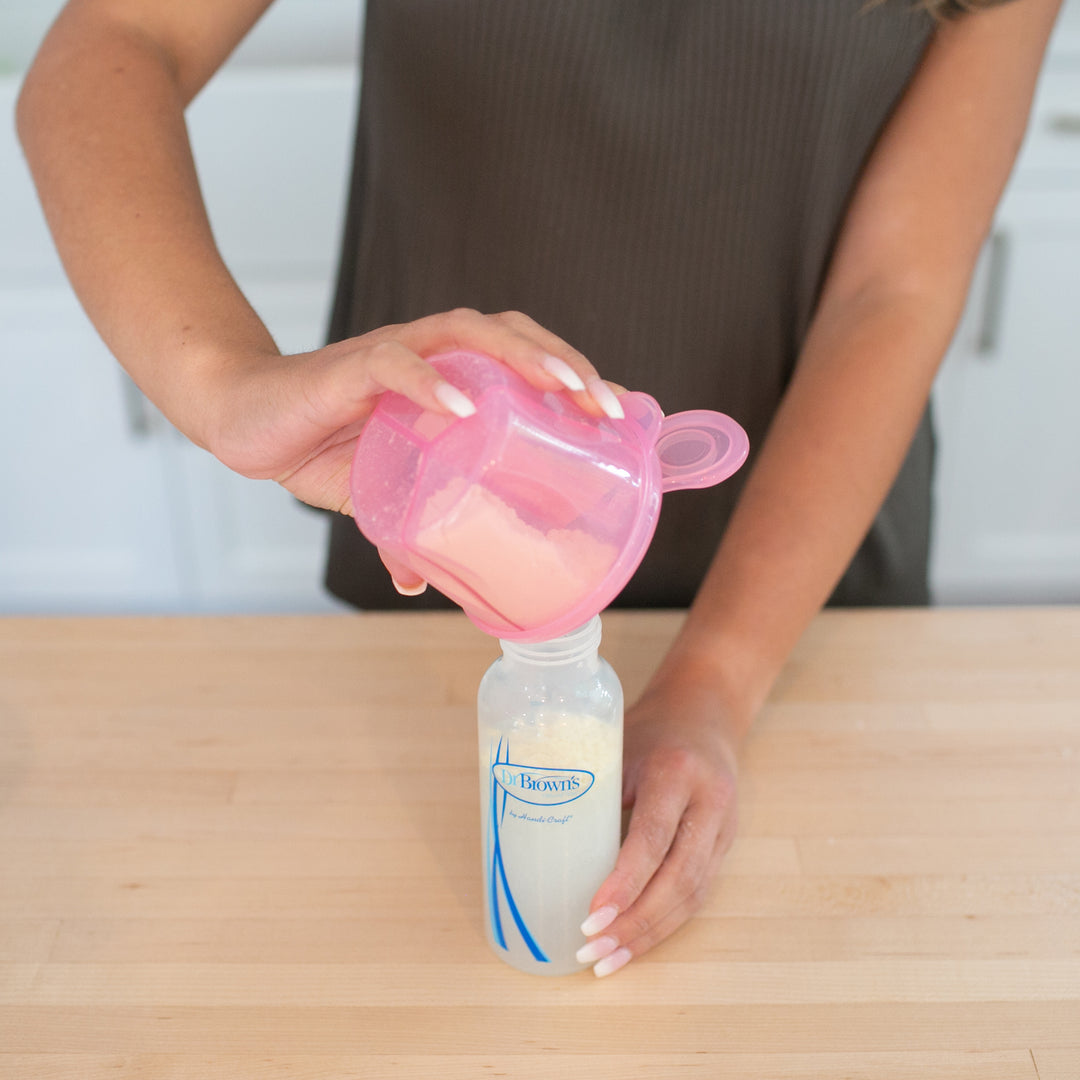 A person carefully pours baby formula from a pink Dr. Brown’s® Baby Formula Dispenser into a baby bottle on a wooden countertop. The transparent bottle with blue and white designs sits against the backdrop of a cozy white kitchen setting.