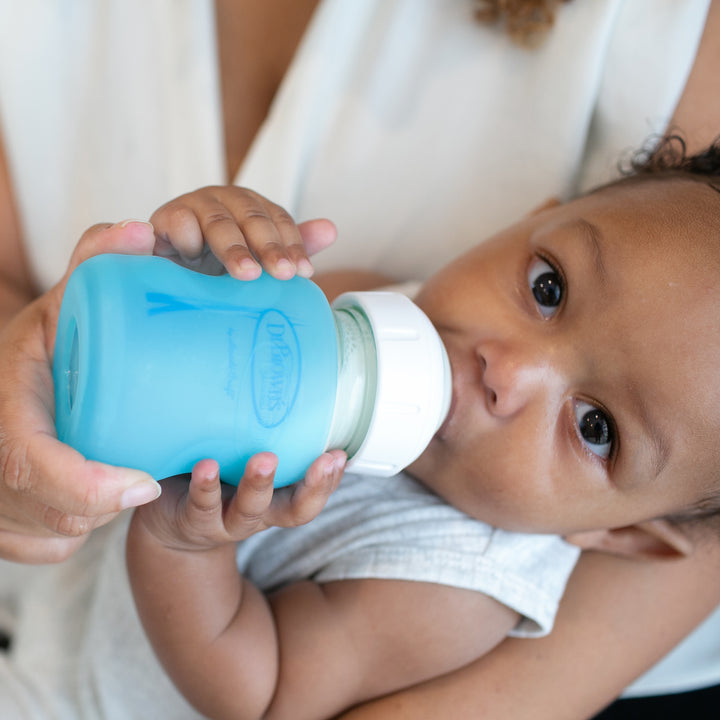 A baby is gently held while drinking from a blue Dr. Brown's Natural Flow® Options+™ Wide-Neck Glass Bottle Silicone Sleeve. The baby looks directly at the camera, dressed in a white outfit and cradled in an adult's arms who is wearing a white shirt.