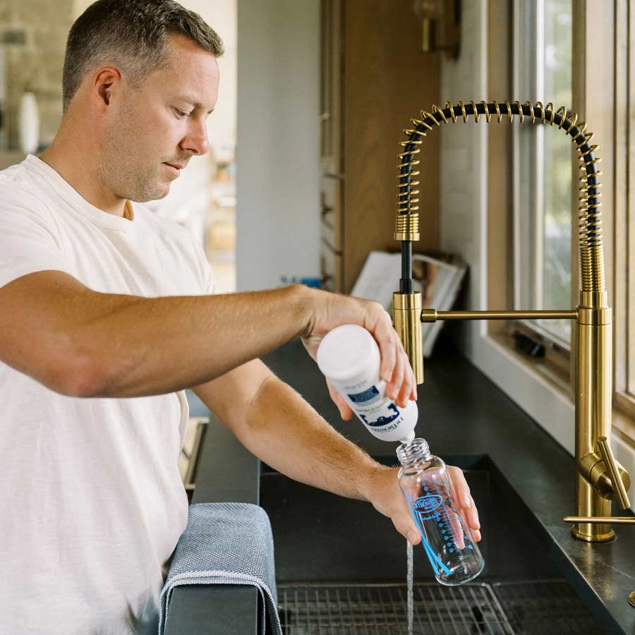 A man wearing a white T-shirt is refilling a water bottle using a filter at the kitchen sink. The sink glistens under the sunlight filtering through the window, made even more radiant by Dr. Brown's™ Bottle & Dish Soap, crafted with natural plant-derived ingredients, and complemented by its brass faucet.