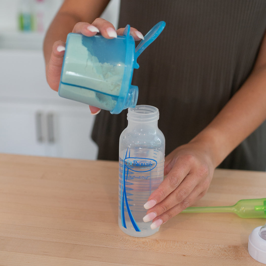 A person is using Dr. Brown’s® Baby Formula Dispenser to pour blue powdered formula into a clear baby bottle placed on a wooden countertop. A green utensil and a bottle cap are nearby, along with other 9 oz bottles. The kitchen backdrop is slightly blurred.