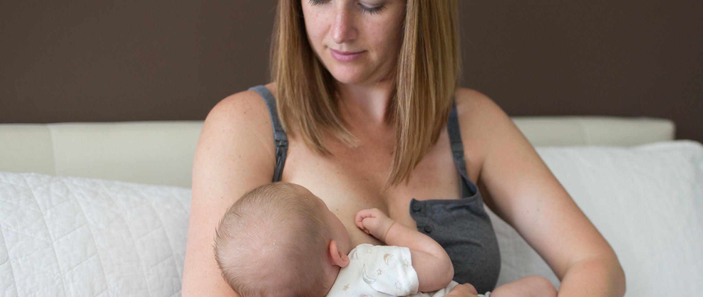 A mother is breastfeeding her baby while sitting on a couch. She is wearing a gray tank top, and the baby is dressed in a white outfit. The background features a brown wall and a white cushion.