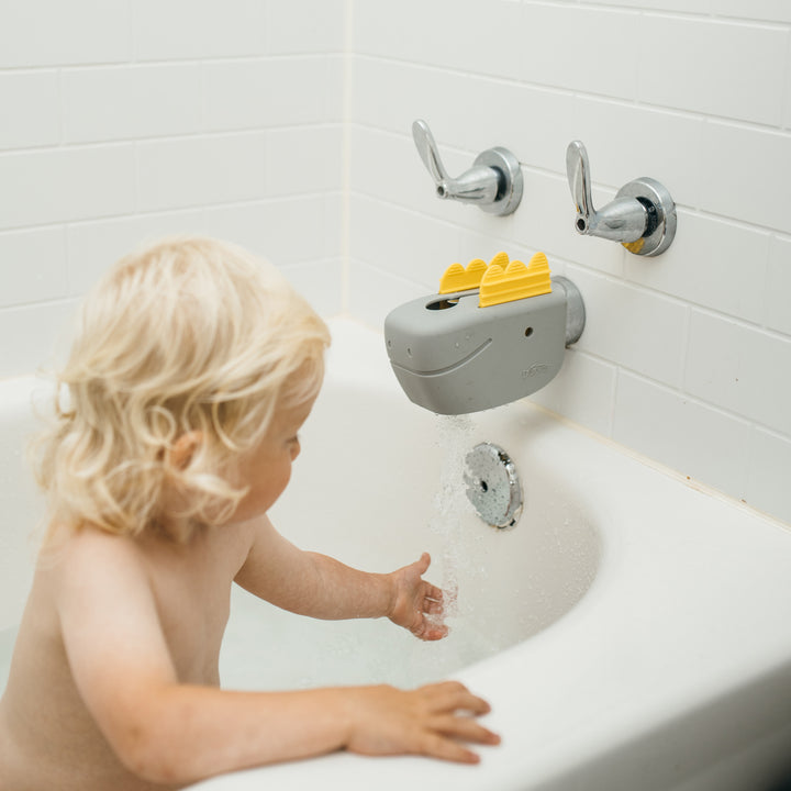 A toddler in a white bathtub enjoys playing with water from a gray, adjustable spout cover designed like a dinosaur. The bathroom features white tiled walls and two silver handles for water control, providing a safe bath time as the child eagerly reaches towards the gentle stream.