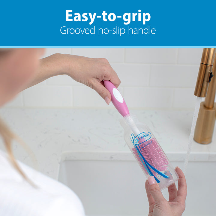 A person is cleaning a baby bottle with Dr. Brown's™ Soft Touch Bottle Brush, which features a pink and white color scheme, under running water. The durable design includes an easy-to-grip, grooved handle for scratch-free scrubbing while a sink and faucet are visible in the background.