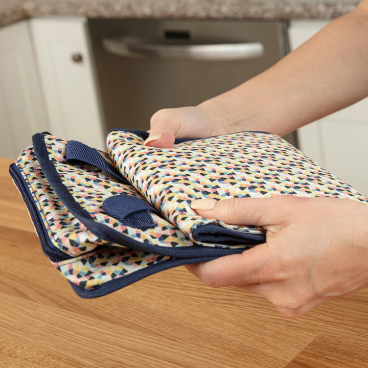 A person holds a set of folded, colorful patterned kitchen towels over a wooden countertop, next to Dr. Brown’s™ Fold & Freeze Bottle Tote. The background features a dishwasher and white cabinets.